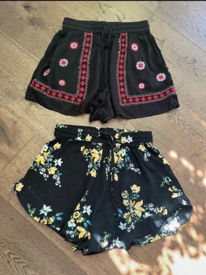 Två svarta shorts från H&M Divided - Säljer två par svarta shorts från H&M Divided i storlek 32. Ena paret har broderade röda och vita detaljer med snörning i midjan, det andra har blommigt mönster i gult, vitt och grönt samt knytband. Båda är tillverkade i mjuk viskos och har loose passform.