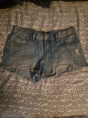 Blå jeansshorts med fransig kant - Säljer ett par blå jeansshorts från 157 med fransig kant och slitna detaljer framtill. Shortsen har klassisk femficksdesign, knapp och dragkedja i midjan samt bakfickor. Perfekta för varma dagar och har en avslappnad loose passform.