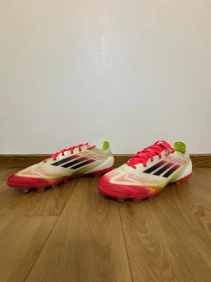 Adidas F50 fotbollsskor vit/röd - Säljer ett par Adidas F50 fotbollsskor med snygg design i vitt, rött och neon-gult. Skorna har svarta Adidas-ränder på sidan, röda snören och platt sula med dobbar. Ovandelen är i syntetmaterial och insidan har neon-gula detaljer. Perfekt för dig som vill sticka ut på planen.