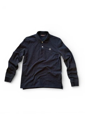 Blå långärmad polotopp från Morris - Säljer en stilren mörkblå långärmad polotopp från Morris med klassisk krage och två knappar framtill. Toppen har ett broderat emblem på bröstet och är tillverkad i mjuk bomullspiké som ger en skön känsla. Perfekt för dig som gillar en clean och tidlös look.