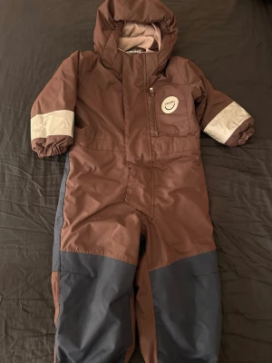 Overall från Viking, strl 98 - Säljer en brun täckoverall från Viking i storlek 98. Overallen har grå förstärkningar på benen, reflexdetaljer på ärmarna och mjukt fleecefoder på insidan. Praktisk dragkedja framtill och elastiska benslut med fotstrappar. Perfekt för kalla dagar ute!