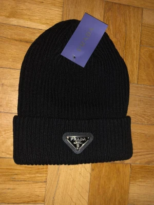 Prada svart mössa - Snygg svart ribbstickad mössa från Prada med uppvikt kant och metallmärke framtill. Mössan har en klassisk form och är tillverkad i ett mjukt stickat material som håller dig varm och stilren.
