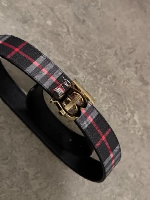 Svart Burberry Bälte med TB logga - Säljer ett snyggt bälte från Burberry i svart, grått och rött med guldfärgad B-spänne framtill. Bältet har ett klassiskt rutmönster och är dekorerad med Burberrys ikoniska ryttare. Perfekt för dig som vill ha en cool och tidlös look.