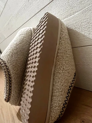 Beige fluffiga tofflor från Duffy  - Mysiga slip-in tofflor från Duffy i beige färg med fluffigt syntetmaterial och dekorativ kant i brunt och vitt. Sulan är tjock och vågformad för extra komfort. Perfekt för chill hemma och håller fötterna varma under kalla dagar.