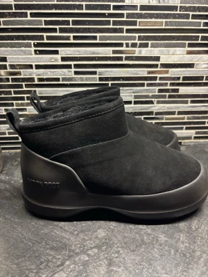 Moon Boot Luna Low Boot Suede svart - Nya Moon Boot Luna Low Boot Suede i svart med rund tå och platt sula. Utsida i mocka och insida med mjuk fuskpäls för extra komfort. Snygg minimalistisk design med dragflik bak och robust yttersula med grepp. Perfekt för kalla dagar.