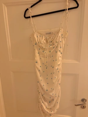 Vit blommig långklänning från Adoore, santorini - Supersöt långklänning från Adoore i vit satin med små blå blommor. Klänningen har smala axelband med knyt, rynkade detaljer och snörning vid bysten. Sval och elegant passform, perfekt för sommarens alla tillfällen.