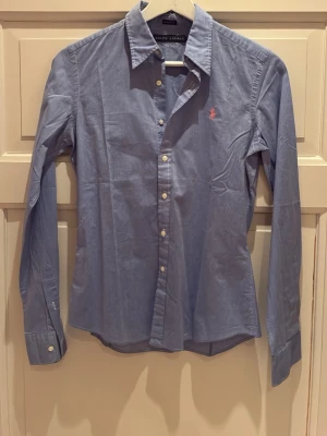 Blå damskjorta från Ralph Lauren - Snygg blå skjorta från Ralph Lauren i skinny fit med klassisk krage och långa ärmar. Skjortan har knappar framtill och en broderad rosa logga på bröstet. Storlek XS. 