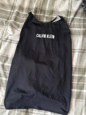 Svart träningslinne från Calvin Klein - Säljer en svart träningstopp från Calvin Klein Performance med vit logga framtill. Toppen har smala axelband och en snygg knytning i ryggen som ger en cool look. Perfekt för träning eller chill dagar. Materialet är mjukt och stretchigt, troligen polyester.