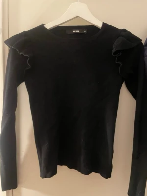 Svart stickad tröja med volang Bik Bok - Svart långärmad stickad tröja från Bik Bok i storlek XS. Tröjan har snygga volangdetaljer på axlarna som ger en cool och trendig look. Passformen är normal och materialet är mjukt och bekvämt. Perfekt för att lyfta din outfit med lite extra stil.
