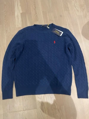 Mörkblå kabelstickad tröja Polo Ralph Lauren - Kabelstickad mörkblå tröja från Polo Ralph Lauren med klassisk röd logga broderad på bröstet. Tröjan har rund halsringning och långa ärmar. Perfekt för dig som gillar stilrena och tidlösa plagg.
