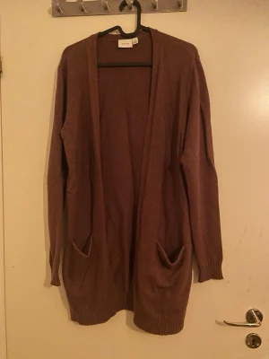 Brun lång kofta från Vila - Säljer en brun lång kofta från Vila med öppet framtill och två stora fickor. Koftan har långa ärmar och ribbade muddar vid ärmslut och nederkant. Perfekt att slänga på sig när det är lite kyligt ute.