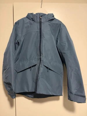 Blå vindj-/regnacka från Everest 134/140 - Blå vindjacka från Everest med huva, hel dragkedja och två stora fickor framtill. Jackan har justerbara ärmslut med kardborre och en reflexdetalj baktill.