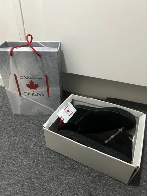 Svarta boots från Canada Snow - Säljer ett par svarta boots från Canada Snow med snygg guldfärgad dragkedja på sidan och rund tå. Skorna är tillverkade i mjuk mocka och har detaljer i skinn samt en platt sula. Perfekta för kalla dagar med stilren look och diskret logga på hälen.