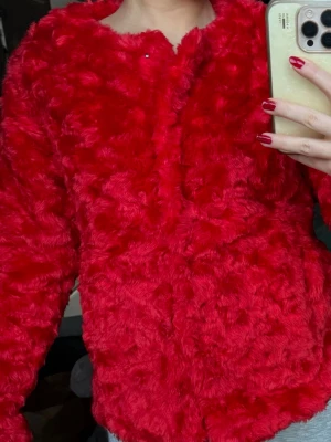 Röd pälsjacka  - Fluffig och supermjuk röd pälsjacka. Jackan har rundad halsringning, lång ärm och stängs med en diskret knapp framtill. Perfekt statement-plagg för dig som vill sticka ut och hålla dig varm. Färgen är intensivt röd och materialet känns lyxigt.