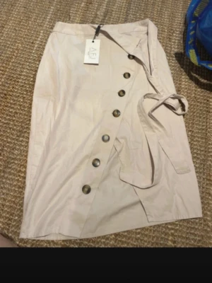 Beige omlottkjol med knappar från AFJ x NA-KD - Säljer en beige omlottkjol från AFJ x NA-KD i storlek 34. Kjolen har snygga knappar framtill och ett matchande bälte i midjan. Den är rak i modellen och passar perfekt till vår och sommar. Materialet är mjukt och känns bekvämt mot huden.