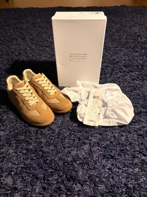 Maison Margiela gats   - Maison Margiela Replica sneakers i beige och ljusbrun färg med snygga paneler i mocka och skinn. Klassisk rund tå, platt sula och snörning framtill. Kommer med originalkartong och dustbag. Perfekt för dig som gillar stilrena och unika sneakers. Det som medföljer är kvitto, box och dustbags. Hör av er vid minsta fundering!