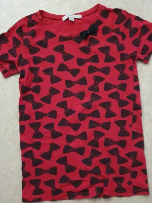 T-shirt - Cool röd t-shirt från MyWear Young med svarta rosetter över hela plagget och en extra tygrosett vid halsen. Rund hals och korta ärmar, perfekt för dig som gillar unika detaljer. Materialet är mjuk bomull som känns skönt mot huden.