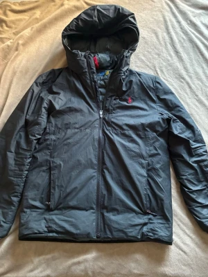 Svart dunjacka från Polo Ralph Lauren - Svart dunjacka från Polo Ralph Lauren med huva och dragkedja framtill. Jackan har en röd broderad logga på bröstet och quiltad insida. Perfekt för kyliga dagar och har en clean, sportig look. Ändast två små håll som knappast syns 