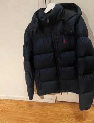 Mörkblå dunjacka från Ralph Lauren - Säljer en mörkblå dunjacka från Ralph Lauren med huva och dragkedja framtill. Jackan har puffad design, två sidofickor med dragkedja och en röd logga på bröstet. Perfekt för kalla dagar och har en riktigt schysst streetstil.