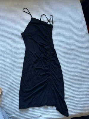 Svart klränning från MAX&Co. - Svart designer klänning från MAX&Co. i storlek S med smala axelband och snygg rynkning längs ena sidan. Har tre axelband som möts i ryggen och sne urringning! Är lite mindre i storleken så är mera en xs/s 