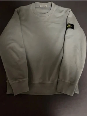 Blå sweatshirt från Stone Island - Snygg ljusblå sweatshirt från Stone Island med klassisk rund hals och ribbade muddar. Tröjan har den ikoniska Stone Island-loggan på vänster ärm. Perfekt för dig som gillar stilrena och enkla plagg med streetkänsla.