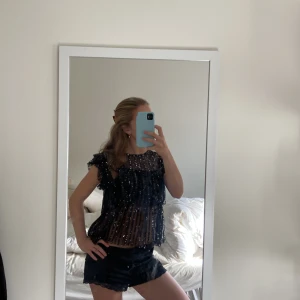 Svart glittrig blus från Gina Tricot - Säljer en svart transparent blus från Gina Tricot med glittriga paljetter över hela toppen. Blusen har volangdetaljer vid axlarna och är ärmlös, vilket ger en cool och festlig vibe. Perfekt för dig som vill sticka ut med lite extra glitter💕