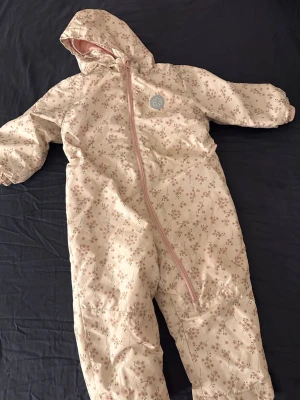 Blommig täckbyxdress från Lupilu, strl 98 - Säljer en supermysig täckbyxdress från Lupilu i storlek 98. Utsidan är ljusbeige med små rosa blommor och har en sned dragkedja framtill. Insidan är fodrad med mjuk rosa fleece som håller värmen. Perfekt för kalla dagar och har huva för extra skydd.
