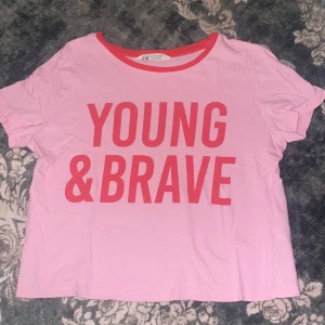 Rosa t-shirt med tryck från H&M - Säljer en rosa t-shirt från H&M med stort tryck 'YOUNG & BRAVE' i rött framtill. T-shirten har rund hals med röd kant och är kortärmad. Magtröja i mjuk bomull.