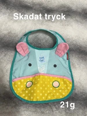 Färgglad haklapp med djurmotiv - Söt haklapp från Skip Hop med ett djurmotiv i blått, gult och rosa. Haklappen har rosa öron, gul prickig nederdel och gröna detaljer. Perfekt för att göra måltider lite roligare.🌸 Skadat tryck efter tvätt.