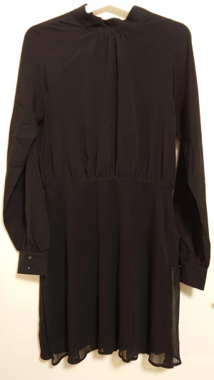 Kort svart klänning storlek Medium 38  - Kort svart klänning med tunna långa ärmar som har knappar vid ärmslut. Dold dragkedja i sidan, v-ringning bak med knytband i nacken. Tyget har ingen stretch. Klänningen är endast provad men lapparna är borttagna. Märke NLY trend Storlek 38 (medium) Längd ca 90 cm Katt finns i hemmet.