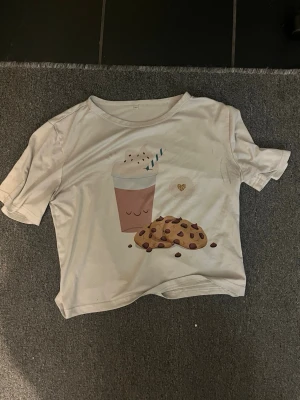 Vit croppad t-shirt med kakmotiv - Söt vit croppad t-shirt med tryck av en kopp varm choklad med vispgrädde och två chocolate chip cookies på framsidan. T-shirten har korta ärmar och en avslappnad passform. Perfekt för dig som gillar gulliga och lekfulla motiv.
