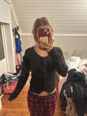 Svart ribbad kofta med knappar - En svart, figurnära kofta med långa ärmar och knäppning framtill. Koftan har en enkel, stilren design och är ribbstickad vilket ger en snygg struktur. Perfekt att slänga över en topp eller t-shirt för en chill look. Omsydd så är väldigt liten i storleken. 