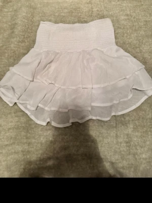 Vit volangkjol med resårmidja - Supersöt vit kortkjol med bred resår i midjan och dubbla volanger. Kjolen är gjord i ett lätt och luftigt tyg, perfekt för varma dagar. Passar perfekt till en avslappnad och trendig stil.