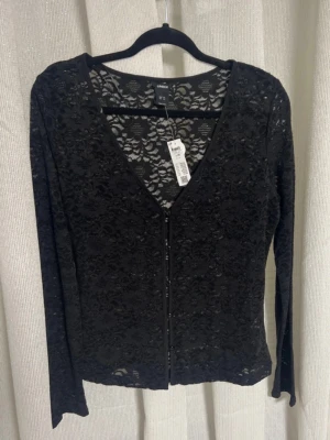 Svart spetsblus med knappar - Trendig svart blus från Lindex  i genombruten spets med blommigt mönster. Blusen har v-ringning, knappar framtill och långa ärmar. Perfekt för dig som vill ha en edgy och stilren look.