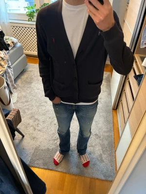 Marinblå cardigan från Ralph Lauren - Pris kan diskuteras!!!! Några frågor är det bara att skriva!!!!!                                 Klassisk marinblå cardigan från Ralph Lauren med V-ringning och knäppning framtill. Tröjan har två fickor och den ikoniska röda logotypen broderad på bröstet. Tillverkad i mjukt material och har lång ärm för en stilren look.