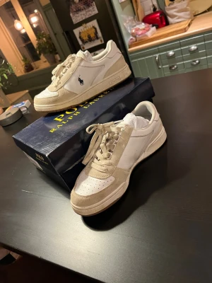 Vita sneakers från Polo Ralph Lauren - Stilrena vita sneakers från Polo Ralph Lauren med beige mockadetaljer och klassisk logga på sidan. Skorna har snörning och en låg profil som passar perfekt till streetwear eller casual outfits. Materialmix av skinn och mocka ger en exklusiv känsla.