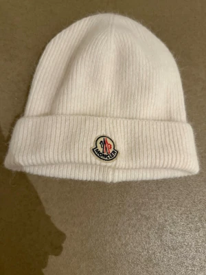Vit ribbstickad mössa från Moncler - Säljer en stilren vit ribbstickad mössa från Moncler med uppvikt kant och klassisk Moncler-logga framtill. Mössan är mjuk och skön, perfekt för kyliga dagar. Passar dig som gillar exklusiva accessoarer och enkel design.