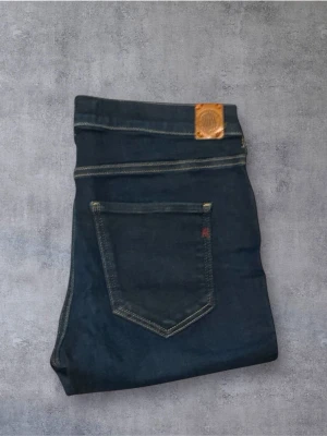 Replay Mörkblå - Snygga mörkblå Replay jeans med klassisk femficksdesign och gul kontrastsöm. Jeansen har en smal passform och är tillverkade i slitstarkt denim med läderpatch bak i midjan. Perfekta för dig som gillar en modern och stilren look.