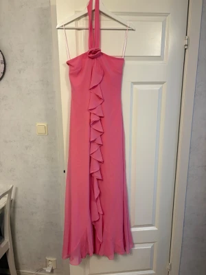Rosa långklänning med volangdetalj - Snygg rosa långklänning med halterneck och volangdetalj framtill. Klänningen har en elegant, flytande passform och är ärmlös. Perfekt för sommarens speciella tillfällen. Har använts en gång och köptes för 1500kr. 