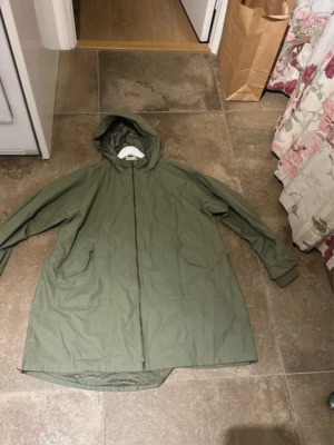 Olivgrön regnjacka med huva - Snygg olivgrön regnjacka med huva och dragkedja framtill. Jackan har två stora fickor och är mycket bra skick, ser knappt använd ut. Den har dekorativa dragkedjor på sidorna och kan spännas åt i midjan med hjälp av resårband. Perfekt för regniga dagar och enkel att matcha med olika outfits. 