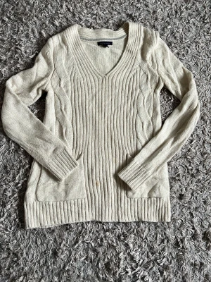 Beige stickad tröja med kabelmönster. Tommy hilfiger. - Snygg beige stickad tröja med V-ringning och kabelstickat mönster längs sidorna. Tröjan har långa ärmar, ribbstickade muddar och två fickor framtill. Perfekt för lager-på-lager och en mysig look.