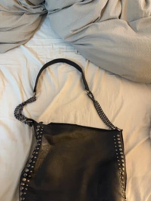 Svart handväska med nitar och kedja - Cool svart handväska i skinn med silverfärgade nitar längs kanterna och en kedjedetalj på axelremmen. Väskan har en mjuk form och dragkedja upptill. Perfekt för dig som gillar en edgy stil.