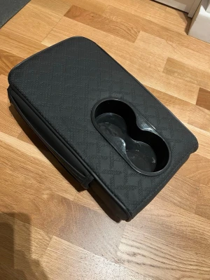 Insats till armstöd i bilen med mugghållare - Den är i svart färg med snyggt mönster. Fack på sidorna och mugghållare uppe. Sätt den på armstödet mellan föraren och passageraren för lite extra höjd och extra förvaring. 