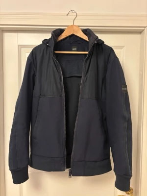 vindjacka Boss - Snygg mörkblå vindjacka från BOSS med huva och dragkedja framtill. Jackan har diskret logga på ärmen, mesh-detaljer på sidorna och ribbade muddar. Perfekt för dig som vill ha en stilren och sportig look.