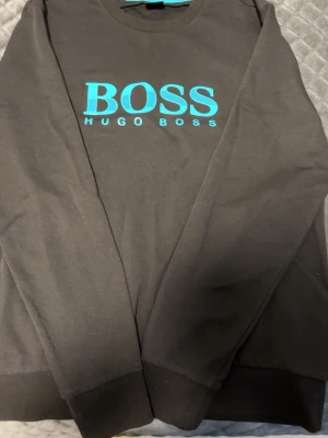 Svart sweatshirt från Hugo Boss - Snygg svart sweatshirt från Hugo Boss med stort turkost BOSS-tryck på bröstet. Tröjan har rund halsringning och ribbade muddar vid ärmslut och nederkant. Perfekt för en stilren och avslappnad look.