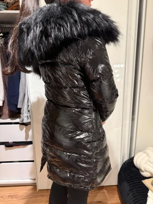 Svart glansig dunjacka med stor pälskrage från femenina - Snygg svart dunjacka med glansig finish och stor, fluffig pälskrage i svart. Jackan har lång passform, quiltade partier och dragkedja framtill. Perfekt för kalla dagar när du vill hålla stilen. Från Femenina Sweden