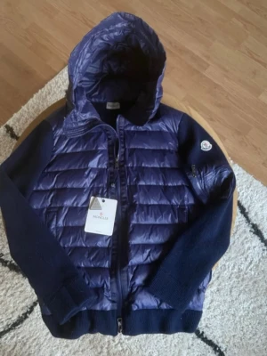 Mörkblå Cardigan från Moncler - Snygg mörkblå dunjacka / cardigan från Moncler med quiltad front och huva. Jackan har stickade ärmar och muddar samt dragkedja framtill. Klassisk Moncler-logga på ärmen och en modern, sportig look. Perfekt för kyliga dagar.