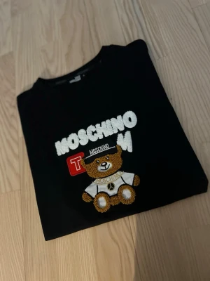 Svart t-shirt med nalle från Moschino - Svart t-shirt från Love Moschino med ett stort broderat nallemotiv och texten MOSCHINO på bröstet. T-shirten har rund hals och korta ärmar. Perfekt statement-plagg för dig som gillar lekfull design och streetstyle.