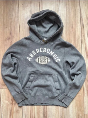 Vintage hoodie från Abercrombie(extremt sällsynt) - Vintage nygg grå hoodie från Abercrombie med stor magficka och tryck framtill med texten 'Abercrombie 92' och en amerikansk fotboll. Hoodien har ribbade muddar och en klassisk huva med metallöljetter. Perfekt för en avslappnad och sportig stil.