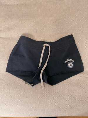 Mörkblå shorts från Hollister - Mörkblå shorts från Hollister med vit snörning i midjan och broderad logga på ena benet. Sköna och avslappnade med kort passform. Midjemått: 36cm, längd: 23 cm 💖